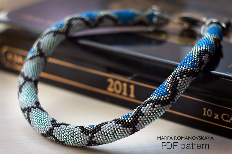 Może przedstawiać: Naszyjnik z koralik&oacute;w w kształcie sk&oacute;ry węża w odcieniach niebieskiego, czarnego, białego i srebrnego. Naszyjnik leży na czarnej książce z nadrukowanym rokiem 2011 na okładce. Pod naszyjnikiem znajduje się napis "MARFA ROMANOVSKAYA PDF pattern".