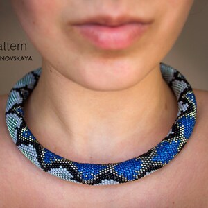 Może przedstawiać: Zbliżenie na naszyjnik z koralik&oacute;w w kolorze niebieskim, czarnym i srebrnym, imitujący sk&oacute;rę węża. Naszyjnik nosi osoba z rozmytym obliczem. W g&oacute;rnej części obrazu widoczny jest tekst "PDF pattern MARFA ROMANOVSKAYA".