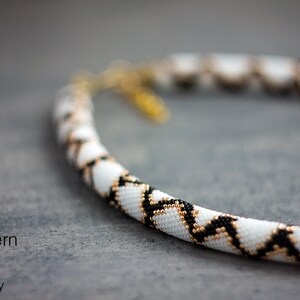 May include: A white, black and gold beaded necklace with a geometric pattern. The necklace is made with small seed beads and is approximately 18 inches long.