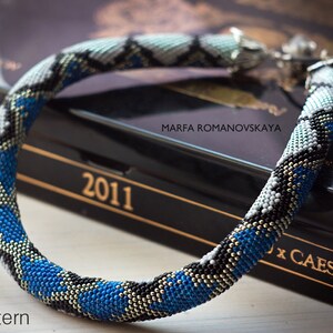 Może przedstawiać: Naszyjnik z niebieskich, białych, czarnych i złotych koralik&oacute;w w geometryczny wz&oacute;r. Naszyjnik leży na czarnym pudełku z napisem "MARFA ROMANOVSKAYA" i "2011".