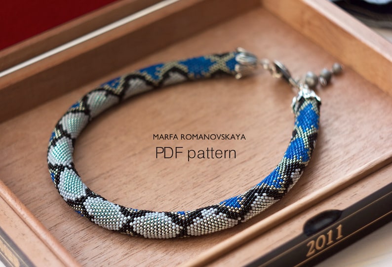 Może przedstawiać: Naszyjnik z niebieskich, czarnych i srebrnych koralik&oacute;w ze srebrnym zapięciem. Naszyjnik znajduje się w drewnianym pudełku z napisem "MARFA ROMANOVSKAYA PDF pattern" i "2011".