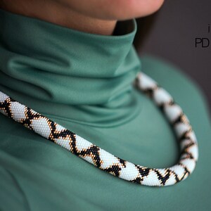 May include: A white, black and gold beaded necklace with a geometric pattern. The necklace is worn by a person wearing a green turtleneck shirt. The text "MRF jewellery PDF pattern" is visible in the top right corner of the image.
