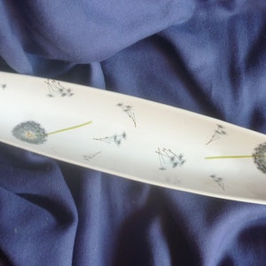 Könnte beinhalten: Weiße ovale Servierplatte mit einem Löwenzahnmuster. Die Platte ist mit einem großen Löwenzahn und mehreren kleineren Löwenzähnen verziert, deren Samen im Wind fliegen.