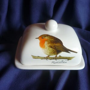Könnte beinhalten: Eine weiße Keramik-Butterdose mit Deckel und kleinem rundem Griff. Die Dose zeigt eine detaillierte Illustration eines Rotkehlchens auf einem Ast. Der Vogel hat eine rote Brust und braune Flügel. Das Wort "Rotkehlchen" ist aufgedruckt.