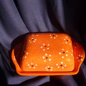 Könnte beinhalten: Eine orangefarbene Butterdose aus Keramik mit Deckel, verziert mit weißen und schwarzen Tupfen. Die Dose hat eine rechteckige Form mit abgerundeten Kanten und einem passenden Tablettsockel. Der Hintergrund ist ein dunkelblauer Stoff.