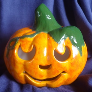 Könnte beinhalten: Ein Keramik-Kürbis mit einem grünen Stiel und einem geschnitzten Jack-o'-Lantern-Gesicht. Der Kürbis ist orangefarben bemalt mit gelben und roten Akzenten.