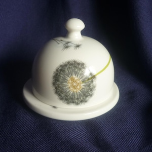 Könnte beinhalten: Eine weiße Keramik-Butterdose mit gewölbtem Deckel und rundem Boden. Der Deckel zeigt ein detailliertes Pusteblumen-Design in Grau- und Gelbtönen mit einem grünen Stiel. Die Dose steht vor einem dunkelblauen Hintergrund.