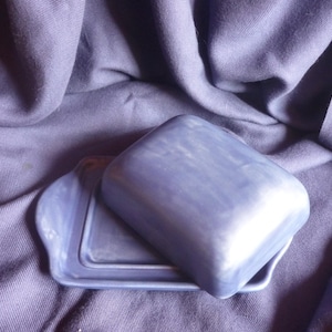 Könnte beinhalten: Eine blaue Keramik-Butterdose mit Deckel, die auf einem passenden Tablett ruht. Die Dose hat eine rechteckige Form mit abgerundeten Kanten. Der Hintergrund ist ein drapierter Stoff.