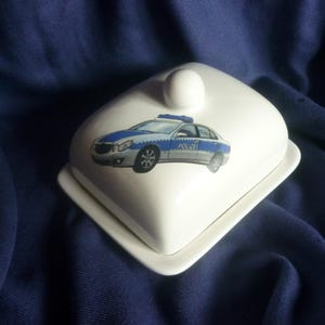 Könnte beinhalten: Eine weiße Butterdose aus Keramik mit einem Deckel, der einen knopfförmigen Griff besitzt. Der Deckel ist mit einer Grafik eines silber-blauen Polizeiautos verziert, auf dessen Seite das Wort POLIZEI steht. Die Dose hat eine rechteckige Basis.
