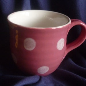 Könnte beinhalten: Eine rosa Keramiktasse mit weißen Punkten. Die Tasse hat eine abgerundete Form und einen Henkel.