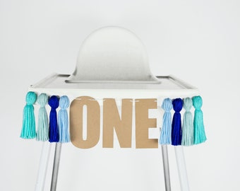 Puede incluir: Una silla alta blanca con una pancarta de cartón marrón que dice "ONE" decorada con borlas de hilo azul y turquesa.