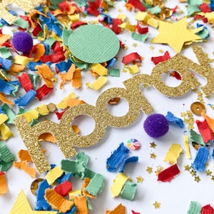 May include: Gold glitter letters spelling "Happy" on a white background with colourful confetti. The confetti is made of paper and includes stars, circles, and strips.