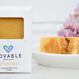 May include: A bar of handcrafted soap, labeled "Bittersweet" by Lovable Body Care, is displayed in a white box with a window. Another bar of soap sits on a white dish. The soap is a light orange color with dark flecks.