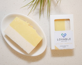May include: A two-tone handcrafted soap bar in a white oval dish, with a white and yellow layered design. The soap is next to a white box with the text "LOVABLE BODY CARE" and "LETS GET TROPICAL HANDCRAFTED SOAP".