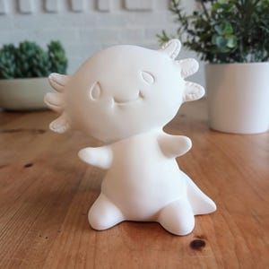 May include: A white ceramic axolotl figurine with a smiling face, small arms, and a tail. The figurine has a textured head with multiple frills. The axolotl is standing on a wooden surface.