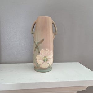 May include: A vintage, light pink ceramic vase with two handles. The vase has a floral design with a white flower and green leaves. The vase is sitting on a white shelf.