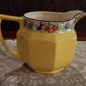 May include: A yellow ceramic pitcher with a floral and fruit design around the top. The pitcher has a octagonal body and a handle.