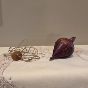 May include: A dark reddish-brown wooden spinning top with a pointed metal tip. Beside the top lies a tan string attached to a small, round, four-hole wooden button. The spinning top has a smooth, rounded body that tapers to a point at the bottom.