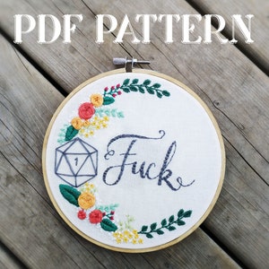 Puede incluir: Arte de bordado en forma de aro con un diseño floral y la palabra "Fuck" en cursiva. Un dado gris de 20 caras con un 1 visible está en el centro del aro.