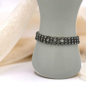May include: A silver-toned bracelet with three rows of sparkling clear rhinestones. The bracelet is displayed on a white, vase-shaped form against a soft, off-white background. The bracelet's design features a repeating pattern of small, round stones.