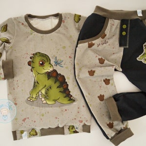 Peut inclure: Une tenue pour bébé en deux pièces avec un imprimé de dinosaure vert. Le haut est un t-shirt à manches longues avec une bordure marron. Le pantalon est noir avec une bordure marron et un patch de dinosaure vert. Le pantalon a un imprimé marron et blanc avec des empreintes de pas de dinosaure.