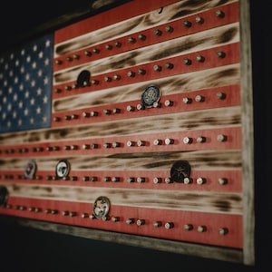 May include: A wooden American flag with red, white, and blue stripes. The flag is mounted on a wall and has a distressed wood finish. There are small round metal objects attached to the flag, some of which have a design on them.