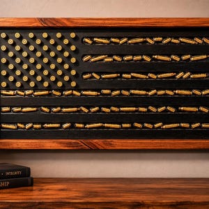 May include: A rectangular wall art piece featuring a wooden frame and a black background. The design mimics the American flag using brass-colored bullet casings arranged in rows to represent the stars and stripes pattern within the frame.