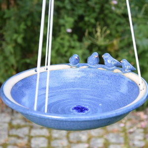 Pode incluir: Um bebedouro para p&aacute;ssaros de cer&acirc;mica azul com quatro pequenos p&aacute;ssaros azuis empoleirados na borda. O bebedouro est&aacute; suspenso por cordas brancas.