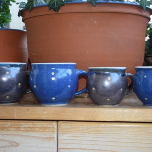 Può includere: Quattro tazze in ceramica blu con pois bianchi. Le tazze sono disposte in fila su una superficie di legno.