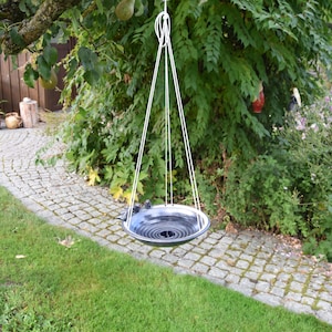 May include: A black and white ceramic bird bath hanging from a white rope in a garden setting. The bird bath is suspended over a stone path.