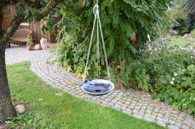 May include: A black and white ceramic bird bath hanging from a white rope in a garden setting. The bird bath is suspended over a stone path.