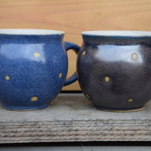 Può includere: Due tazze in ceramica con motivi a pois. La tazza a sinistra è blu con pois gialli. La tazza a destra è grigio scuro con pois gialli.