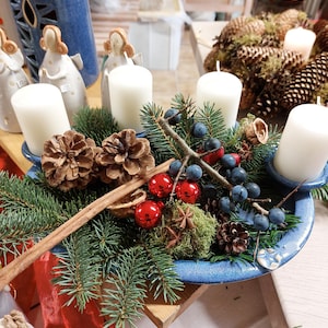 Op de afbeelding: Een feestelijk middelpunt met vier witte kaarsen op een blauwe keramische schaal. De opstelling omvat dennenappels, dennentakken, rode ornamenten, blauwe bessen en kaneelstokjes, waardoor een seizoensgebonden vakantiedisplay ontstaat.