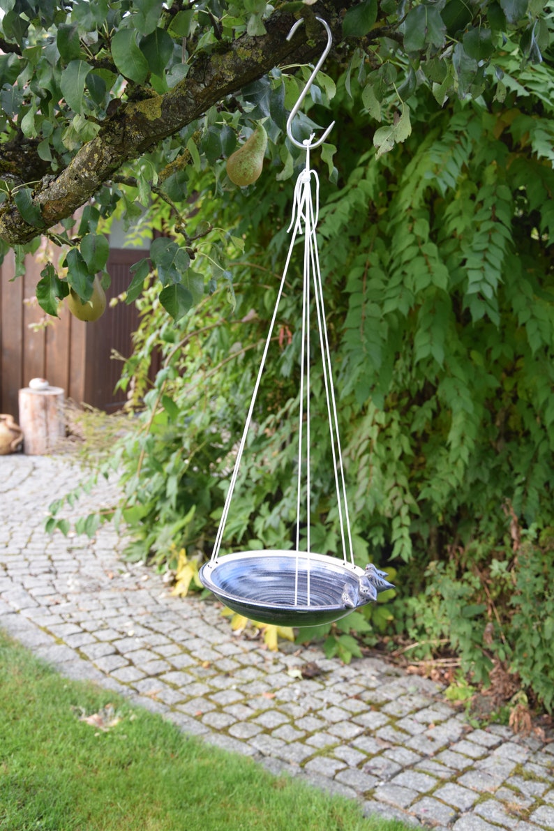 May include: A black and white ceramic bird bath hanging from a tree branch. The bath is suspended by a white rope and has a metal hook.