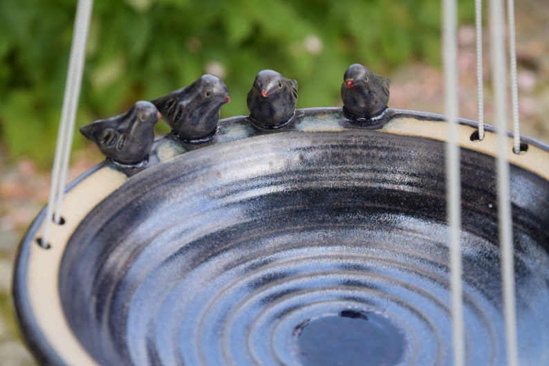 May include: A ceramic bird bath with a blue and white glaze. Four small black birds are perched on the rim of the bath. The bath is suspended from a white cord.