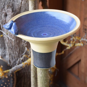Op de afbeelding: Een keramisch vogelbad met een diepblauwe binnenkant en een lichtbeige rand. Twee kleine blauwe vogelfiguren zitten op de rand. De basis is contrasterend zwart en beige. Het vogelbad is op een tak gemonteerd.