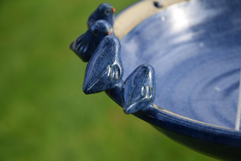 Pode incluir: Um bebedouro para p&aacute;ssaros de cer&acirc;mica azul com quatro pequenos p&aacute;ssaros azuis empoleirados na borda. O bebedouro est&aacute; cheio de &aacute;gua e tem um esmalte branco.