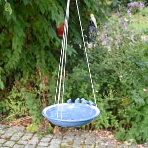 Pode incluir: Um bebedouro para p&aacute;ssaros de cer&acirc;mica azul com uma borda branca pendurado em uma corda em um jardim. O bebedouro tem quatro pequenos p&aacute;ssaros azuis empoleirados na borda.