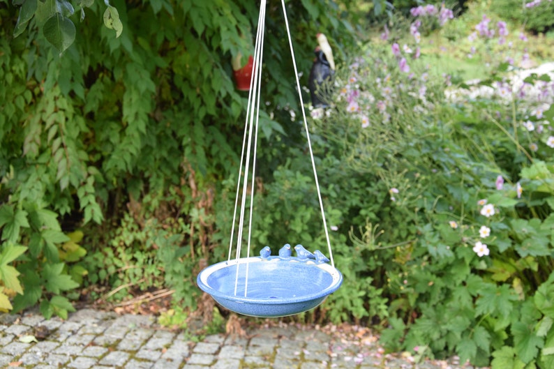 Pode incluir: Um bebedouro para p&aacute;ssaros de cer&acirc;mica azul com uma borda branca pendurado em uma corda em um jardim. O bebedouro tem quatro pequenos p&aacute;ssaros azuis empoleirados na borda.