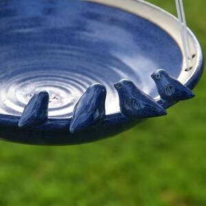 Pode incluir: Um bebedouro para p&aacute;ssaros de cer&acirc;mica azul com quatro pequenos p&aacute;ssaros azuis empoleirados na borda. O bebedouro est&aacute; pendurado em um cord&atilde;o branco.