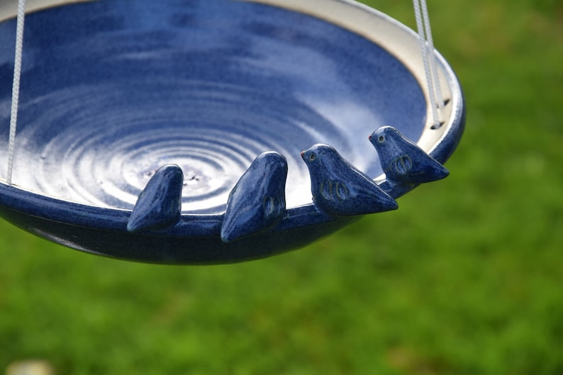 Pode incluir: Um bebedouro para p&aacute;ssaros de cer&acirc;mica azul com quatro pequenos p&aacute;ssaros azuis empoleirados na borda. O bebedouro est&aacute; pendurado em um cord&atilde;o branco.