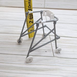 May include: A miniature gray walker with white wheels and a white seat. A yellow measuring tape is positioned behind the walker, showing a height of approximately 2 inches. The walker is designed with a sturdy, geometric frame.