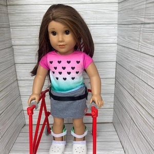 Peut inclure: Une poupée portant une chemise rose et verte avec des cœurs noirs, une jupe grise et des chaussures blanches utilise un déambulateur rouge avec des roues noires.