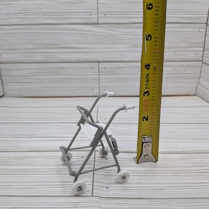 May include: A miniature gray walker with white wheels and handles. The walker has a small white seat. A yellow tape measure is next to the walker, showing the walker's height is approximately 3 inches.