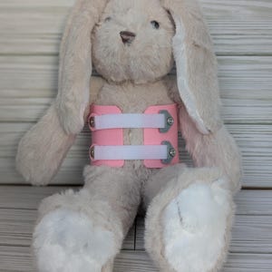 May include: A plush bunny rabbit in a light beige color, wearing a pink brace around its torso. The brace has white straps and silver hardware. The bunny has long ears and white paws.