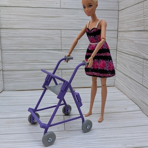 May include: A doll in a black and pink floral dress stands next to a purple walker with gray wheels. The doll has blonde hair styled in a bun. The walker has a gray seat and handles.