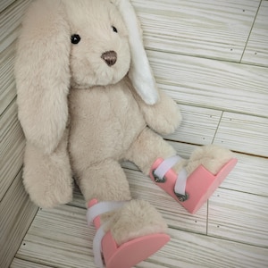 Può includere: Un coniglio di peluche beige con lunghe orecchie floppy e punte bianche, che indossa stivali ortopedici rosa e bianchi.