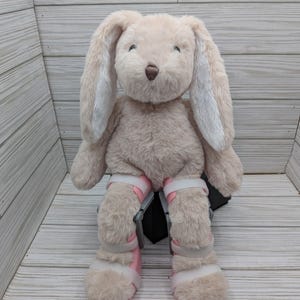 May include: A plush beige bunny with long ears and a brown nose. The bunny wears pink and white leg braces. The toy is positioned against a light wood-grain background.