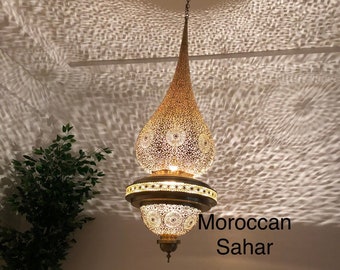 May include: A gold Moroccan Sahar pendant light fixture with intricate geometric patterns. The light fixture is hanging from a ceiling and casting a decorative shadow on the wall.