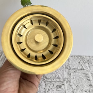 May include: A gold-colored metal sink strainer with a circular design and multiple small holes. The strainer has a raised center with a small button.